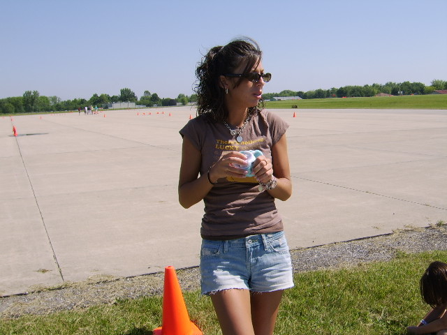 In the image, a young woman stands centrally in the foreground, holding a can of soft drink. She is dressed in casual attire, including a brown t-shirt, jean shorts, and sunglasses. The woman is smiling, and the open can suggests she's enjoying a drink. In the background, beyond the large grassy area adjacent to a sealed cement surface, there's a hint of trees and an expansive sky that stretches to meet the horizon. Further back, smaller figures can be discerned near the concrete, possibly other people at an event or gathering. The setting appears to be a park or open outdoor space.