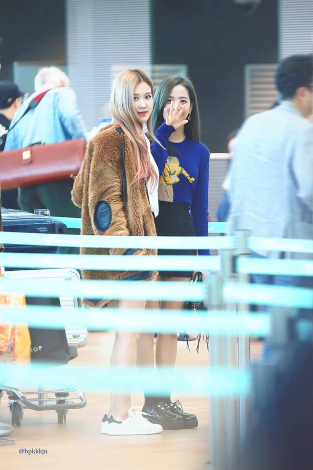 A woman sitting on top of a wooden bench - This image captures a moment at an airport terminal. In the center, two women with long hair stand close together, engaged in what appears to be a phone call. They are both casually dressed, with one wearing a long brown coat and the other in a blue shirt with a graphic design. They are positioned against a backdrop of a bustling terminal filled with passengers, a security barrier and large pieces of luggage. The terminal has a contemporary design with a combination of blue and white color scheme.