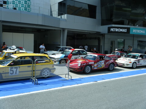 Pistonheads Afos Sepang - The image features a vibrant gathering of race cars lined up next to a track. The cars, varying in design and color, are parked in a staging area, suggesting they are awaiting a race or practice session. In the background, a building with a sign indicating 'PETRONAS' looms, adding context to the location. People, likely participants or staff, can be seen in the vicinity of the cars, contributing to the dynamic atmosphere of the scene. The overall arrangement of the cars indicates a sense of order and readiness for the upcoming event.