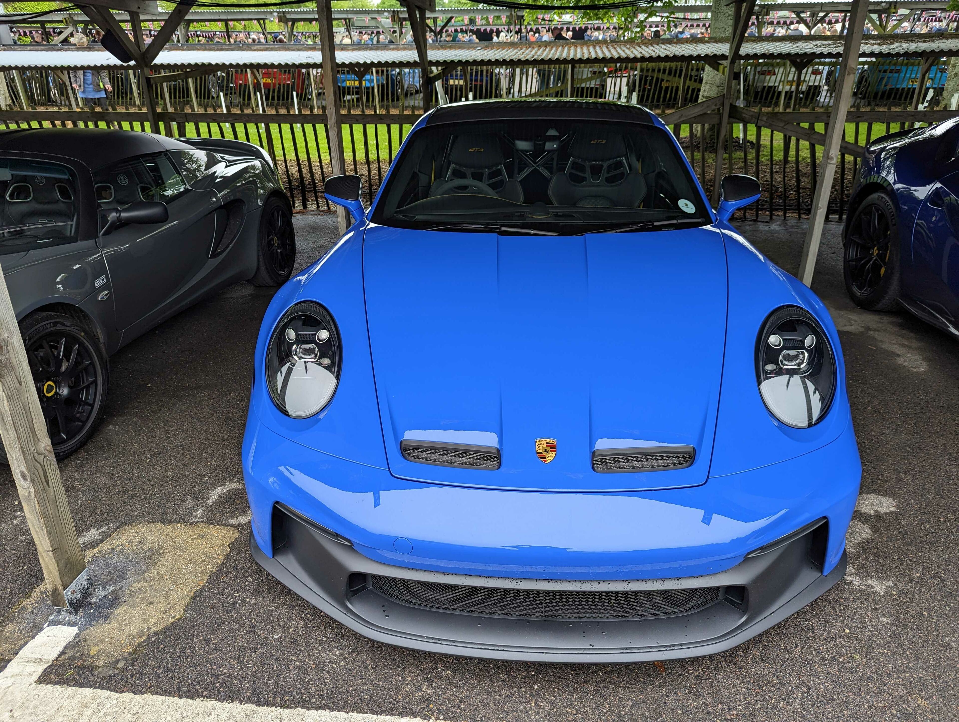 2023 Breakfast Clubs - Page 2 - Goodwood Events - PistonHeads UK - The image shows a blue sports car parked in a parking lot with other cars visible in the background. There is a person partially visible on the left side of the image, and the setting suggests an outdoor event or gathering. The car has a sleek design with visible headlights and alloy wheels. The overall impression is that of a high-end vehicle at a public event.