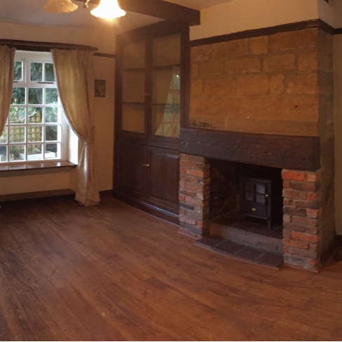 Show me your wood burner before and after pics  - Page 4 - Homes, Gardens and DIY - PistonHeads - The image shows an interior space, specifically a room with a fireplace and wooden paneling. The walls are painted in a light color, and there is a large window on the left side of the image that lets in natural light. A small chandelier hangs from the center of the ceiling. A white curtain partially covers the window, partially allowing in daylight. The room features a wooden floor and is furnished with a wooden cabinet against one wall. There is no visible text or distinctive branding in the image. The style of the photograph seems candid, perhaps taken to document the condition of the property or for real estate purposes.