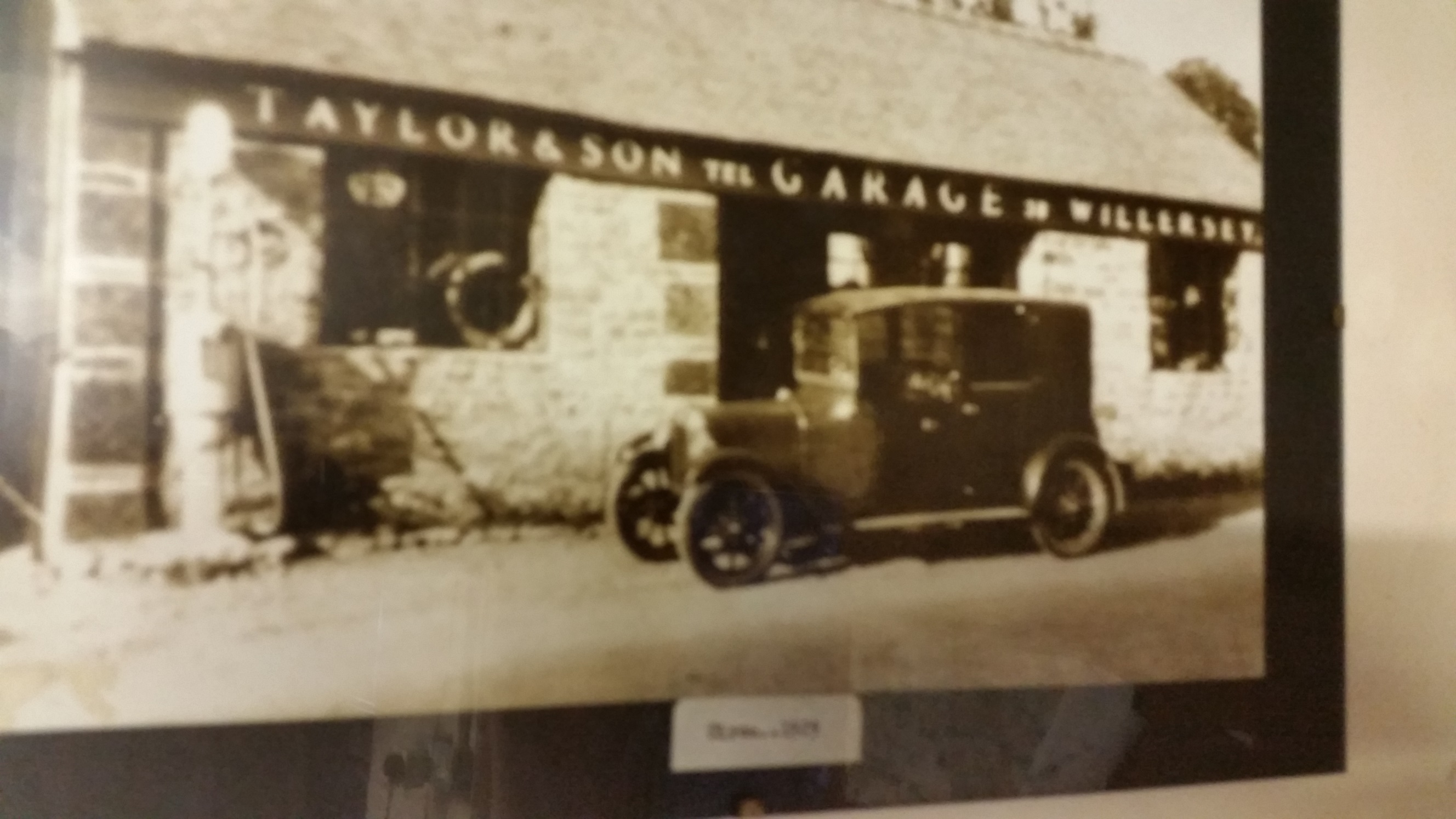 The Humer Unbeam Interesting Filling Stations Thread - Page 38 - General Gassing - PistonHeads - This is a black and white photograph showing the exterior of a building with the sign "TAYLORSON'S MOTOR CYCLE MANUFACTURERS" at the top. There's a vintage automobile parked in front of the building, which appears to be an older model given the make and model designation "WILLERS". In the corner of the frame, there's a reflection suggesting it is taken in a room with a glass enclosure. There is also a piece of paper at the bottom right of the image with the text "April, 2023", which indicates that this image might have been taken or accessed in April 2023.