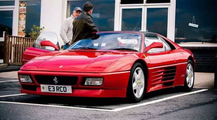 Ferrari 348 chit chat - Page 1 - Ferrari V8 - PistonHeads - The image shows a vibrant red Ferrari parked on the left side of a blacktop parking lot. The car is facing towards the right side of the image where there's a building with windows, and a blue car can be seen in the background. Two men are standing near the Ferrari; the man closest to the car is looking towards the interior of it, while the other person is also present in the vicinity. On the hood of the Ferrari, a license plate with the letters "E3RCO" is clearly visible.