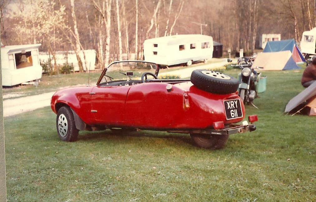 Berkeley  - Page 1 - Classic Cars and Yesterday's Heroes - PistonHeads - This image captures a scene in a camping area. The star of the image is a classic red sports car, prominently parked on a grassy field and taking up a sizeable portion of the frame. The car features a distinctive split window design, reminiscent of certain styles from the 1950's or 1960's. In the immediate surroundings, there are several recreational vehicles, including a caravan, a trailer, and motorcycles, suggesting the presence of a campsite nearby. On the right side of the image, there's a camper with a blue and white awning, providing a pop of color against the green backdrop. The location is situated on a lush green field, providing a serene and picturesque environment for the campers.