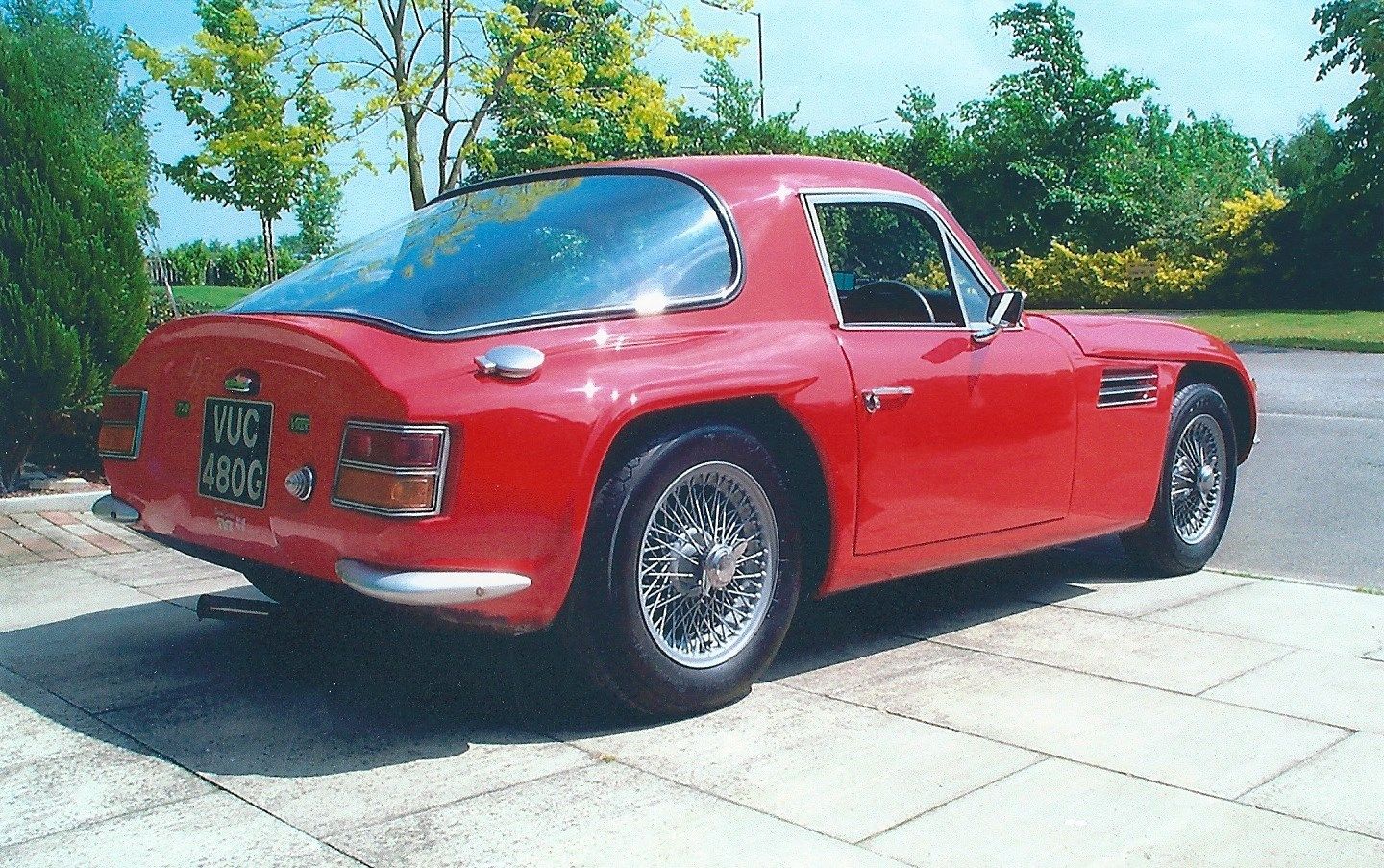 Early TVR Pictures - Page 40 - Classics - PistonHeads - The image showcases a classic red sports car parked on a brick pavement. The vehicle features a small, hardtop design with a long hood, two doors, and a short rear deck. The car has been well maintained, as indicated by its two-tone paint job and the presence of a shiny chrome bumper. A distinctive triangular-shaped badge is visible on the hood, suggesting the brand of the car. The background consists of tree branches and a framed, panoramic sunroof, adding to the overall aesthetic of the vehicle.