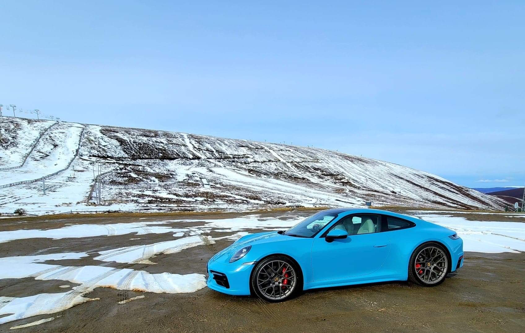 A picture a day... Porsche - Page 136 - Porsche General - PistonHeads UK - The image depicts a striking blue Porsche sports car parked on a gravel surface amidst snow-covered mountains. The vehicle, with its sleek design and distinctive red brake calipers, is the central focus of the composition. The sky above is clear, providing a serene backdrop to the scene. In the distance, the vast expanse of the snowy mountains stretches out, creating a sense of isolation and tranquility. The overall mood of the image is one of calm and beauty, capturing a moment of quiet solitude in a breathtaking natural landscape.