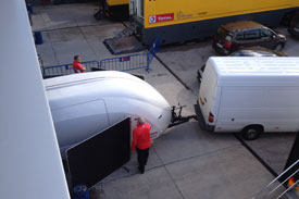 Pistonheads - The image shows a busy parking lot with many vehicles. A white Mercedes-Benz truck is in the process of maneuvering and is in close proximity to a black car. A person is standing outside the truck, who appears to be unloading or performing some activity with the adjacent large white trailer. In the background, another large truck is visible. The setting appears to be an industrial area with storage or warehouse facilities nearby. The image suggests a loading or unloading scene in a commercial area.