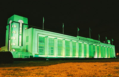 Iconic buildings - That make you go....... - Page 7 - The Lounge - PistonHeads - The image features a large, modern building at night, illuminated predominantly in green and white hues, possibly due to neon or colored lighting. The building has a symmetrical facade with multiple levels, and a tall clock tower is central to the architecture, served by flags mounted on poles at the top of the structure. The grounds surrounding the building are lush and green, and no people are visible, emphasizing the tranquility and possibly the solitude of the location. There's a sense of grandeur and scale to the building within the context of the surrounding landscape. The photograph is taken from a distance, placing the building and its clock tower in the center, with the clock tower being the tallest structure in the frame.