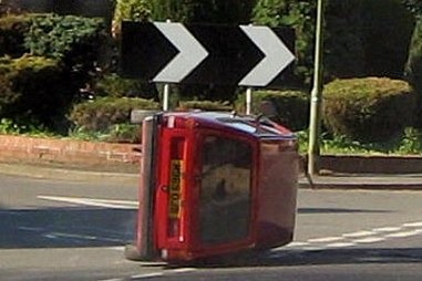 Seconds Pistonheads Endulge - The image shows a scene of a red car, which has flipped on its side and is jutting out onto a road at a street corner. The car is stationary, obstructing the road, possibly caused by an accident. Above it, there are two road direction signs, white with black arrows, indicating a right turn. The photo captures the situation from the back of the car, with a clear sky in the background. It seems to be a quiet, safe neighborhood, but the red car is an unexpected hazard on the road.