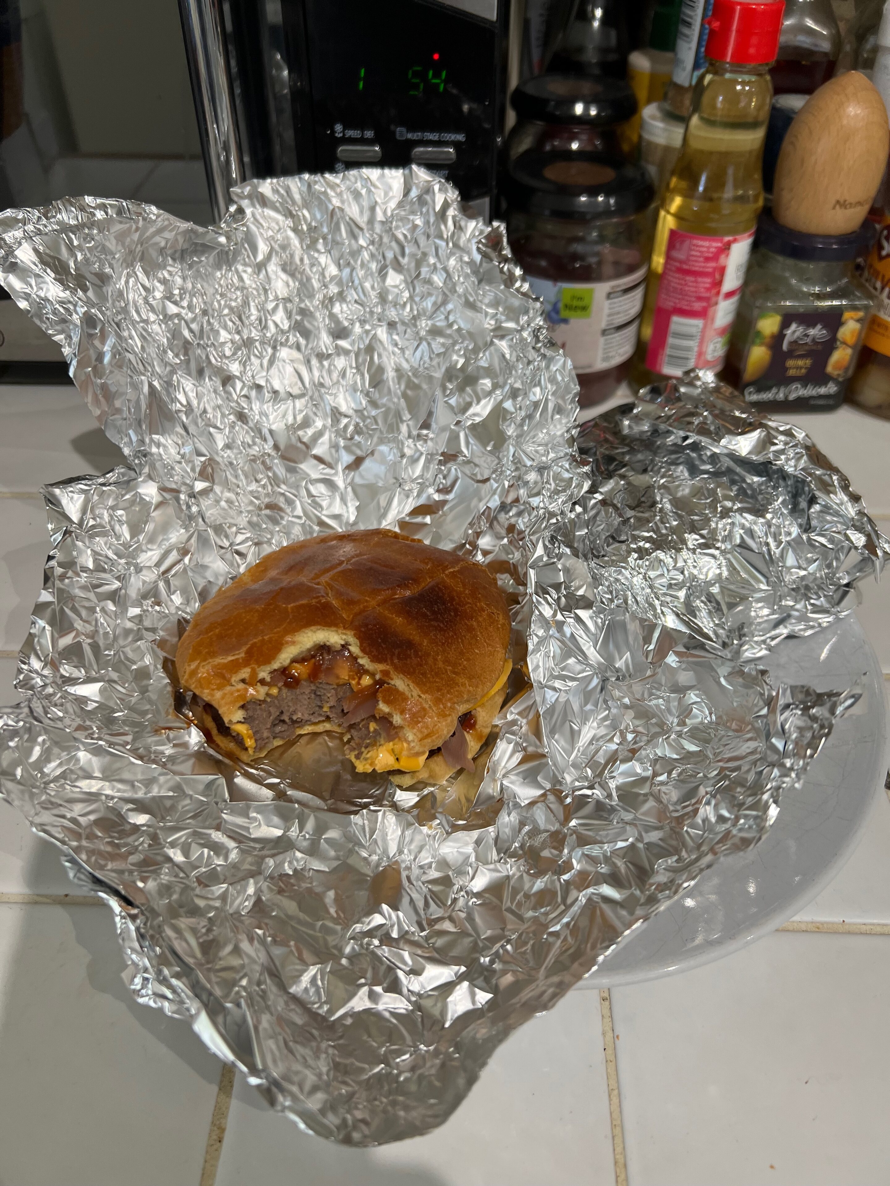 Pistonheads - The image shows a sandwich, possibly a hamburger or burrito, resting on top of some aluminum foil in a white plastic container. The container is placed on a kitchen counter, with various items like bottles and jars scattered around the background. The food is wrapped in foil, indicating it might be a takeout order or prepared for transportation. The counter itself appears to be part of a domestic kitchen setting.