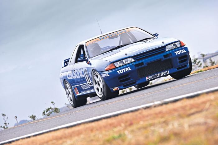 A large truck with a car on the back - Pistonheads - The image features a dark blue Acura Integra car with a prominent grille, driving on a road. The vehicle has a sporty design, indicated by its aerodynamic bodywork. The word "TOTAL" is prominently displayed on the front of the car, suggesting a sponsorship or association with the Total oil company. The background is overcast, which could imply an earlier time of day or simply an overcast sky. The scene captures the car in motion, conveying a sense of speed.