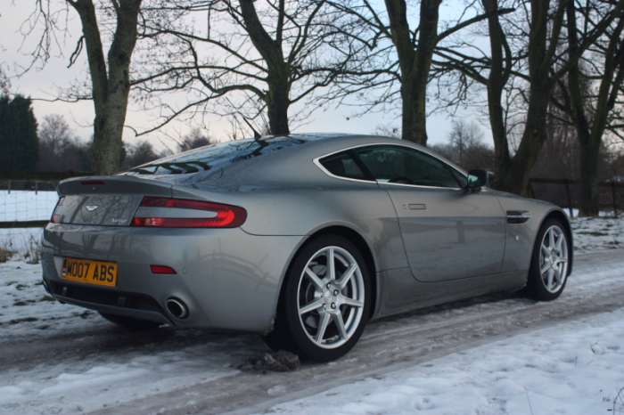 Spot the Difference - Page 1 - Aston Martin - PistonHeads - The image features a sleek, silver Aston Martin sports car parked on a snowy road. The car's sleek design and the snowy background suggest a serene winter setting. The vehicle's license plate is clearly visible, with the registration reading "AM070 ABS." The Aston Martin emblem, a renowned symbol of luxury and performance, is clearly visible on the car's grille, and the wheels are covered in light dustings from the recent snowfall. The sun is low on the horizon, casting a blue hue over the scene and casting long shadows that enhance the car's presence on the road.