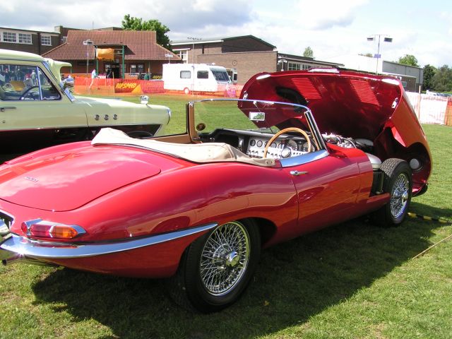 Pistonheads - The image shows a vintage sportscar, possibly a Lotus or a similar model, with an open hood, parked on a grassy field. The car is painted a shiny red and features a classic design with a visible bonnet opening revealing the engine. It is positioned between two other cars, one in front, which appear to be more recent models, and one in the background. The grass field suggests this could be an outdoor car show or exhibition.