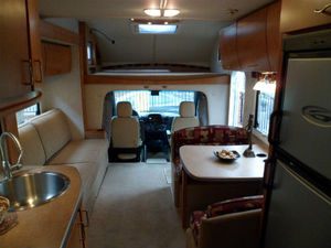 Selling RV? Best Way - Page 1 - Tents, Caravans & Motorhomes - PistonHeads - This image features the interior of a motorhome or camper, showcasing its size and layout. There's a forward-facing window for visibility and a unique ceiling compartment, possibly for storage. The living area is furnished with a seating area that includes a couch and chairs, which are complemented by a small TV mounted on the wall. On one side, a well-equipped small kitchen area is visible with seating, a sink, and cupboard space, completing the mobile living space.