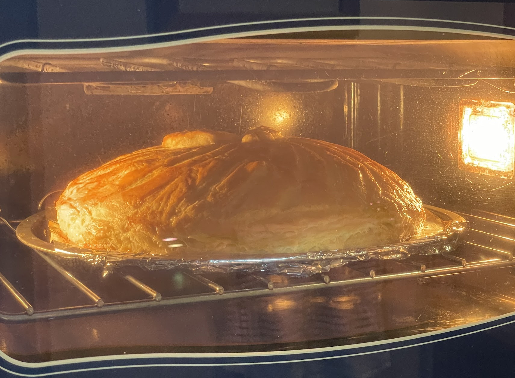 The image shows a large, golden-brown pastry baking in an oven. The oven is open with the light on, illuminating the food inside. The pastry appears to be a pie or tart due to its flat surface and crusty edge, which are common characteristics of these baked goods. The interior of the oven has a clean appearance, and there's no visible text in the image. The style of the photo is straightforward, capturing the cooking process without any additional context or decoration.