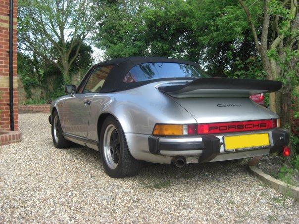 Porsche 911 3.2 - Page 1 - Readers' Cars - PistonHeads - The image shows a silver Porsche 911 Targa car with a distinctive targa top, parked on a gravel driveway. The setting is outdoors, surrounded by lush green trees and bushes indicating it might be late summer or early spring. The sky is overcast, implying a gloomy or possibly a rainy day. The license plate at the back of the car is yellow, which might be unusual depending on the country where the photo was taken. The rear view of the car emphasizes its sleek and classic design.