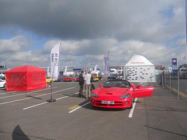 A red car is parked in a parking lot - Pistonheads - The image shows an outdoor area during the daytime. In the foreground, a vivid red sports car is parked on the concrete, facing to the side of the image. There are several people in the background, some of whom are standing underneath a blue and white canopy, likely engaged in conversations. The surrounding environment includes more cars parked in the background, parked under another red and white shelter. A few flags are visible, possibly indicating that this is a special event or gathering. A white tent is set up further back, providing a contrast to the red shelters. The sky overhead is overcast, suggesting a cool day.