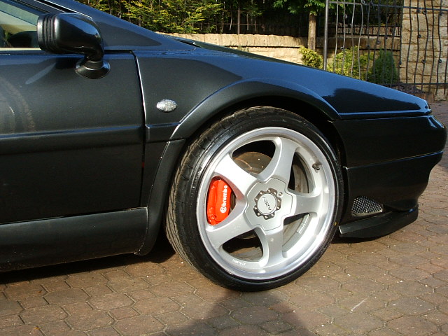 Esprit Pistonheads Market Alloys - The image shows a close-up view of the front wheel of a dark-colored sports car. The wheel, which features orange calipers and silver alloys, is off the ground, mounted on a tire that is slightly turned towards the right. The pavement has a patterned brick design, which is visible in the foreground and background of the image. There are also glimpses of foliage in the background, suggesting that the car is parked near a garden or park. There is no text visible in the image. The car's design and the brick pavement give the image an urban or suburban feel.