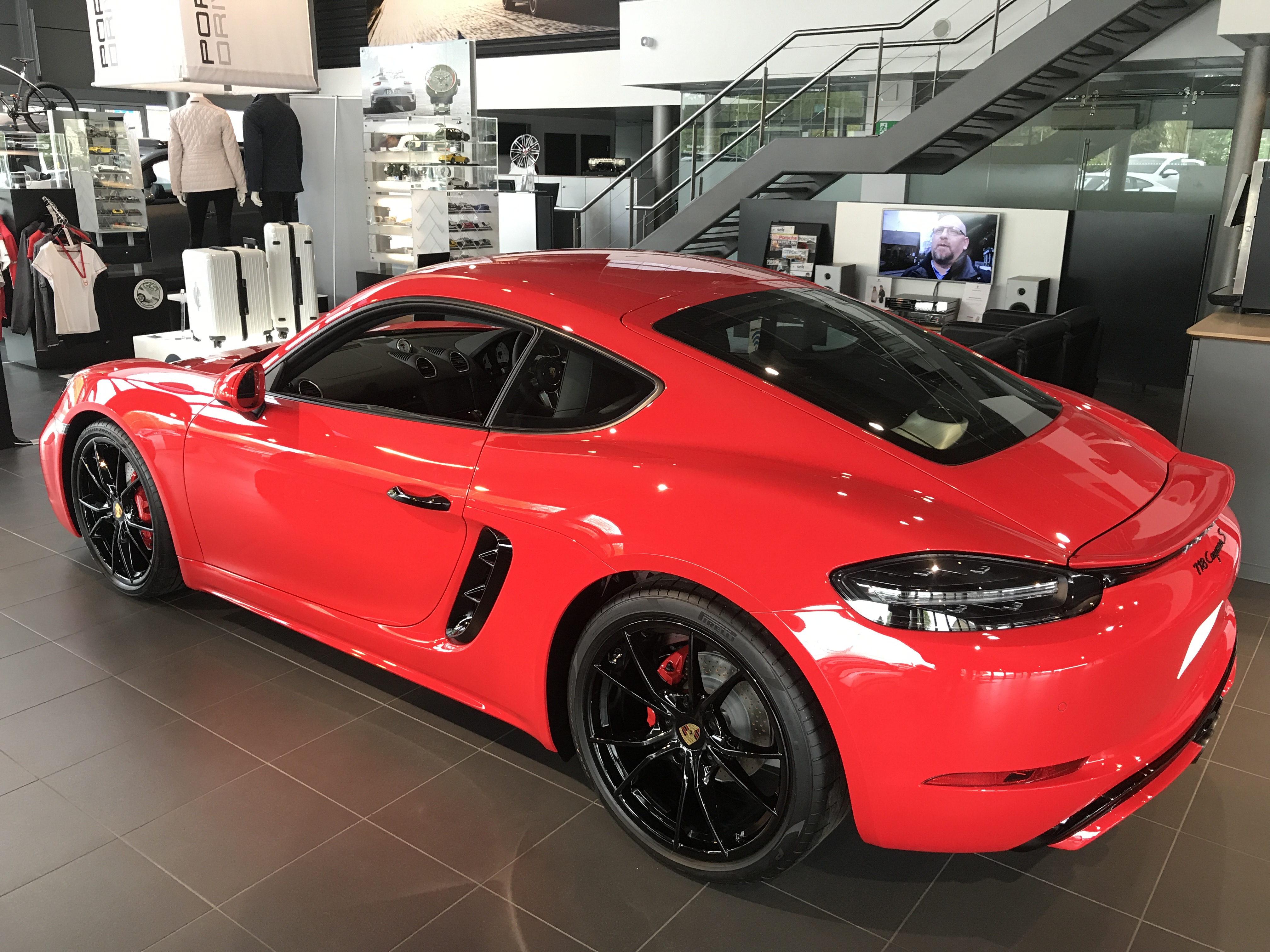 718 Cayman Spec & Colours- what have you gone for? - Page 59 - Boxster/Cayman - PistonHeads - The image showcases a striking red Porsche sports car in a showroom setting. The car's glossy finish reflects the surrounding environment, with black tires mounted and painted accents highlighting its aerodynamic design. The interior can be seen through the large windows that allow for ample natural light to illuminate the vehicle. In the background, the showroom displays escalators, other vehicles, and a variety of accessories, creating an atmosphere of luxury and excitement.