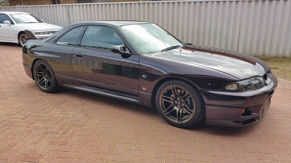 Dark purple black metallic paint examples - Page 3 - General Gassing - PistonHeads - The image shows a sports car parked on a brick pavement next to a white fence in a driveway. The car is black and glossy with a spoiler on its hood. It features black wheels and body details, emphasizing its sleek design. The vehicle's side and part of the front are clearly visible, revealing the side door handle and the driver's headlight. The car is positioned at an angle, highlighting its length and aerodynamic flair.