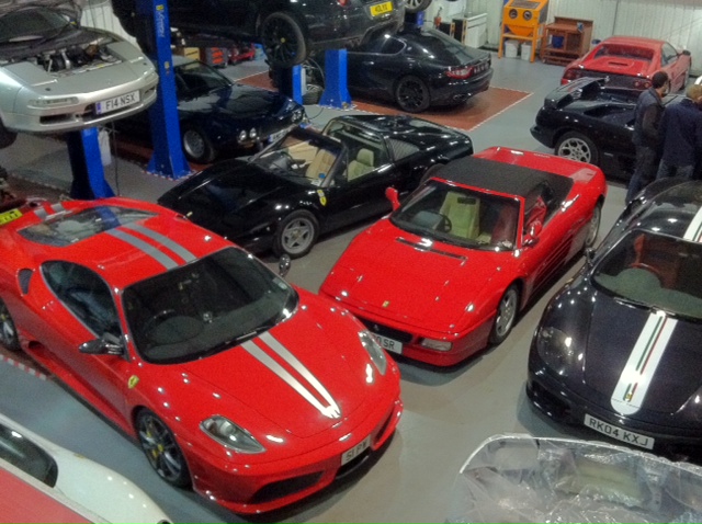 The GT Cafe Gallery - post your meeting photos here - Page 1 - North West - PistonHeads - The image shows a spacious garage filled with a variety of luxury sports cars. These cars are of different colors and models, including bright red, sleek black, and elegant silver. The vehicles are parked or parked on a display of red bricks, which adds a touch of contrast to the garage's monochromatic tones. In the background, there is a person present, likely a staff member or visitor to the garage. The overhead lighting illuminates the scene, casting a warm glow over the cars and enhancing their reflective surfaces.