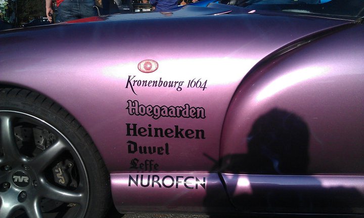 Random TVR pics - Page 68 - General TVR Stuff & Gossip - PistonHeads - The image shows the driver's side door on a car. The surface of the car's paint has several colorful and fancy labels. These labels feature the text "KROENBURG 1604" and "HEINEKEN," along with other words, indicating sponsorship or branding by these companies. There's an intricate image of a car wheel with the word "NUROFEN" written on it. The background outside of the car is blurred, drawing the focus to the decorative labels on the car's surface.