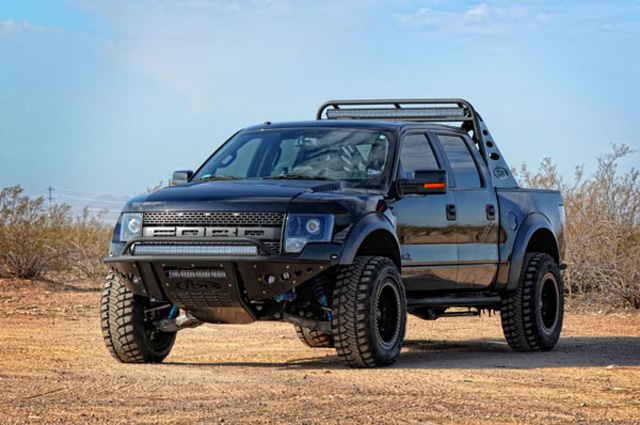 Pistonheads - This image shows a black truck with robust off-road capabilities. The truck is parked on a dry, sandy terrain, surrounded by sparse vegetation. Its design is rugged and utilitarian, with a prominent front grille, large flat-fender arches, and a modified suspension with increased ground clearance. The vehicle features chunky off-road tires, a high-rise off-road bumper with integrated fog lights, and a substantial roof rack. The sky above is blue, indicating fair weather conditions, which is appropriate for such a vehicle due to its adventurous nature.