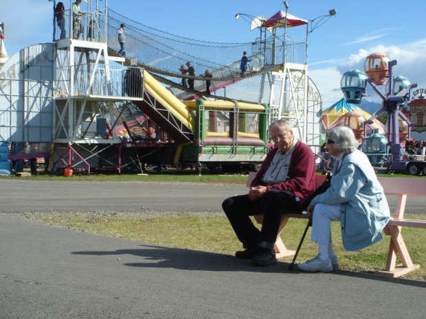 Aston Martin World! - Page 1 - Aston Martin - PistonHeads - The image features an elderly couple sitting on a park bench in front of a vibrant amusement park with a high-energy environment. They appear to be conversing and enjoying the lively view.