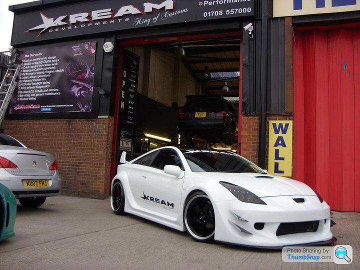 Badly modified cars thread - Page 134 - General Gassing - PistonHeads - The image displays a white sports car parked in front of a red garage door with the word "CREAM" written above it. On the car, there is a name or logo "KREAM" along with other text, and a photographer's name or watermark at the bottom right of the image. In the background, there are additional vehicles, and a sign with "PERFORMANCE" and a phone number visible.