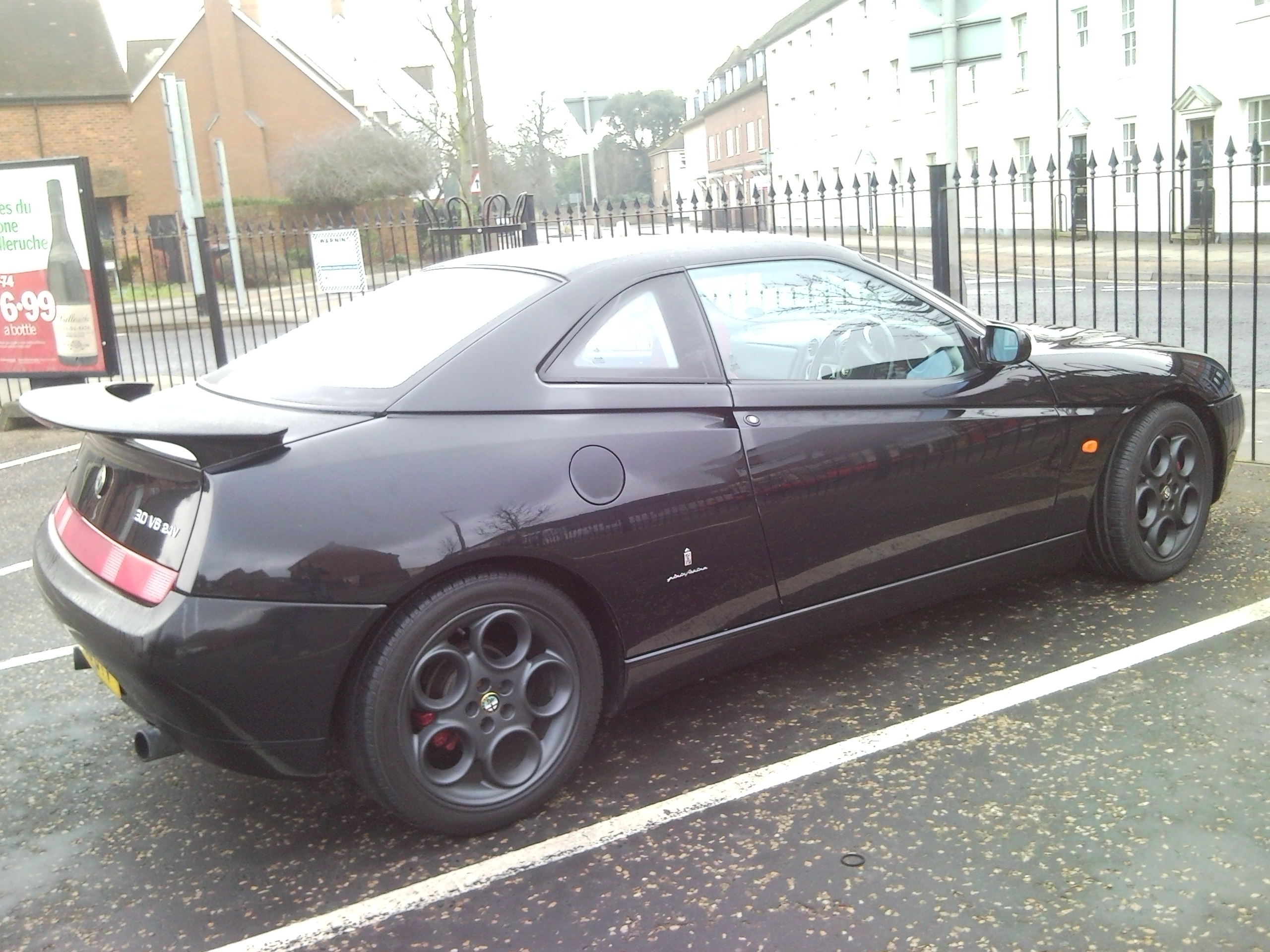 Let's see your Alfa Romeos! - Page 26 - Alfa Romeo, Fiat & Lancia - PistonHeads - The image depicts a parked black sports car, which seems to be a coupe model, given the design of the rear and the side windows. It appears to have black rims with a special design, maybe customized or from a sports package. The car is parked in a lot, with the bicycle lane visible in the image to the left, suggesting that there might be dedicated bike lanes nearby. The car is positioned next to a black fence and there are high-rise buildings in the background, indicating that this scene might be in an urban area.