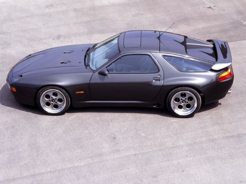 RE: Porsche 928: Catch it while you can - Page 3 - General Gassing - PistonHeads - The image shows a dark gray sports car parked on a street with its hood up, revealing the engine area. The car has a rear-wheel-drive (RWD) configuration, indicated by the vents on the rear quarter panel. It features alloy wheels and a retractable rear spoiler. The photograph captures the car from a three-quarter angle with the front end facing towards the camera's left.