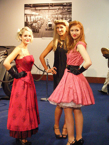 Pistonheads - This is a color photograph featuring three young women standing together. They are dressed in vintage-style dresses appropriate for an upscale event or vintage-themed gathering. The dresses appear to be richly textured. Two of the women have hairstyles with bouffant arrangements, and one has smooth, straight hair. They all have makeup and may be posing for an event or a special occasion. The background is not clearly discernible but provides a context in which the individuals are standing, suggesting an indoor event or exhibition.