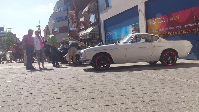 Camberley Town Centre car show - Page 1 - Thames Valley & Surrey - PistonHeads - The image captures a scene on a city street, where a vintage red and white convertible is parked. People are gathered around the car, showing interest in it. Building advertisements can be seen in the background, lending a vibrant urban atmosphere to the scene. There are two cars parked on the street: one is the vintage red and white convertible, while the other is a compact black car. It appears to be a sunny day, as the street is bustling with activity and the buildings tower in the background.