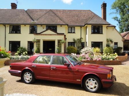 Probate Value - Bentley Turbo R - Page 1 - Rolls Royce & Bentley - PistonHeads - The image depicts a red, two-door luxury car parked in front of a large, yellow multi-story house with a white trim. The car has a gleaming finish and white alloy wheels. The house features a long driveway and a well-maintained front lawn with flowering plants and shrubs. The sky is clear with a few clouds scattered in the distance, suggesting a sunny day.