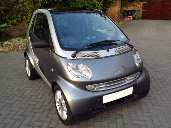 New winter beast! - Page 1 - North West - PistonHeads - This image features a compact Smart Car parked in a driveway. The car has a sleek, modern design with a shiny metallic body, reflecting some of its surroundings. The car's back windshield is visible, and it bears a white license plate. The setting is a suburban residential area, with a brick driveway and greenery in the background.