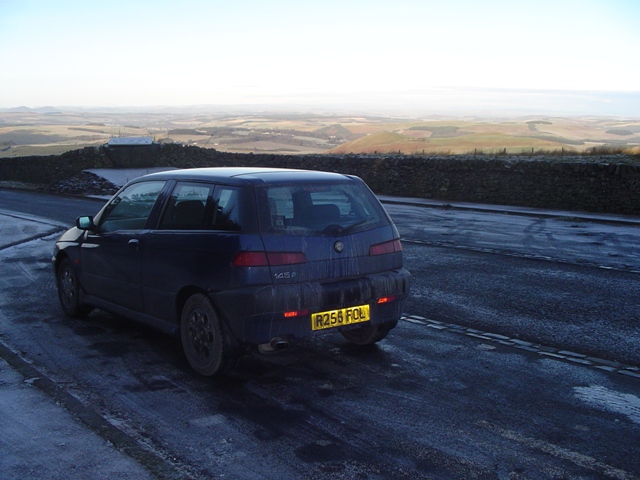 Pistonheads - The image shows a dark blue four-door car parked on a road. The surroundings appear to be a mountainous landscape with a barren and rocky terrain. The car is situated on the right side of the road, as indicated by the license plate that reads "245 FOL." The sky is clear, suggesting it might be a cold day given the brackish appearance of the water bodies around the car. The road itself shows signs of previous traffic, with streaks and spots along its length.