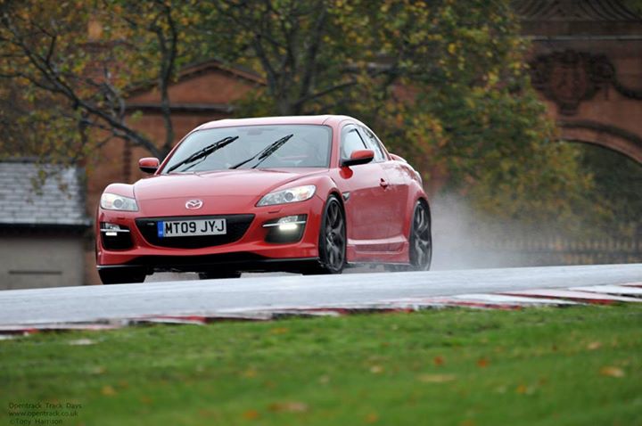 Mazda RX8 Track Car, "The Kraken" - Page 1 - Readers' Cars - PistonHeads - The image captures an intense moment on a rainy day. A shiny red sports car is driving through water splashing from the road, suggesting high speed. The vehicle is positioned in the foreground and takes up a significant portion of the frame, emphasizing its prominence. Behind the car, the environment is equally dramatic. Dark, possibly red brick buildings rise up, forming an archway or possibly a tunnel entrance on the right side of the image. A lush green grassy area stretches towards the watermark bearing the text "SATURDAY TIMES." The overall scene is dynamic and vibrant, showcasing the car's performance in challenging weather conditions.