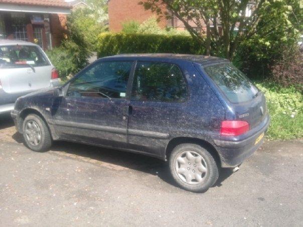 Pistonheads - This image shows a used, small hatchback car parked on a gravel driveway. The car's body exhibits streaks of dirt and mud, indicating recent wet conditions or rough driving. There are signs of wear and age on its paint surface. The environment around the car includes a brick building in the background and a tree filled with green leaves to the right. The car has a main color of dark blue, with a silver trim and a bright red taillight.