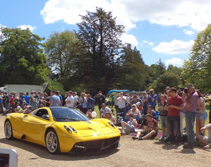 Wilton house  - Page 1 - South West - PistonHeads - This is a Technicolor landscape featuring a bright yellow sports car as the centerpiece. The car is parked on a gravel area, facing towards the viewer, and is surrounded by a crowd of people who appear to be enthusiasts or spectators. The people are spread out around the car, some seated on chairs, while others stand tall taking pictures or simply admiring the vehicle. The background reveals an expansive field, which is flanked by lush green trees, contributing to a serene and picturesque atmosphere.