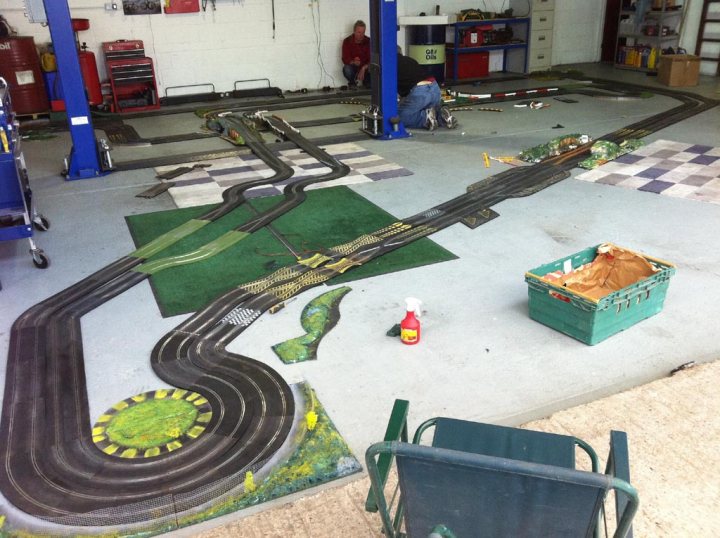 Van Pistonheads Transit Scalextric Challenge - The image shows a large toy race track set up indoors, taking up a significant portion of the space. It is a carbon-copy setup, with smooth curves and straight sections designed to resemble a real racetrack. There are various toy cars on the track, ready for a race. On the right side of the image, there's a blue chair facing away from the viewer. On the sparse floor, there are a couple of small engineering components, including a syrup bottle and a pair of shoes. There's a glimpse of a workbench with tools in the background, suggesting an area for repair or modification of the track or cars.