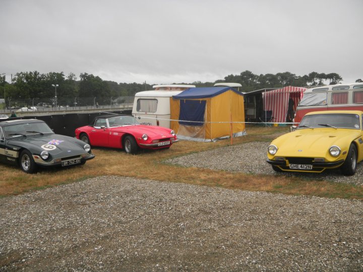 le mans classic  - Page 1 - Classics - PistonHeads - The image showcases a gravel area where three vintage cars are parked close together. The cars exhibit an array of vibrant colors including red, yellow, and black. These vehicles - possibly sports cars - are positioned in a unique formation within the frame. The background features a white car, adding a contrasting touch to the scene, and an orange tent set up in the distance. The sky above is filled with clouds, suggesting an overcast day.