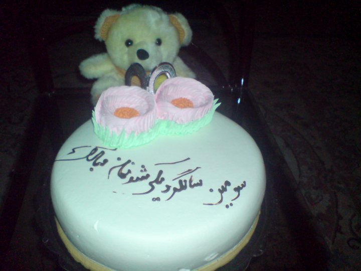 Bear Cake Toy - The image shows a table with a cake and a teddy bear. The cake is round and has pink frosting with feet details. It also has Arabic writing on it. The teddy bear is sitting next to the cake. Its fur appears to be white, with orange ears. The bear has a playful expression on its face. The setting suggests a celebration or party. The lighting is soft, and the focus is on the cake and the teddy bear, making them the main aspects of the image. The overall mood seems to be festive or celebratory.