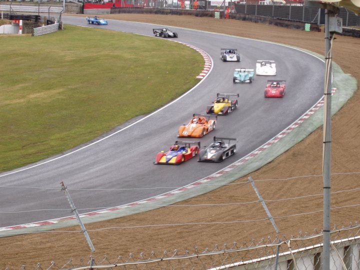 BRSCC Meeting Brands Hatch 11-06-2011 - Page 1 - UK Club Motorsport - PistonHeads - The image depicts an exhilarating race taking place on a winding, gray racetrack. Multiple race cars are engaged in a competitive race around a corner. The cars are a mix of colors such as yellow, red, and blue, and they are different types indicating a variety of engines or categories. The racetrack is bordered by a grass infield, adding to the lush green landscape surrounding the track. Above the track, there's a clear blue sky, suggesting a beautiful day at the racetrack.