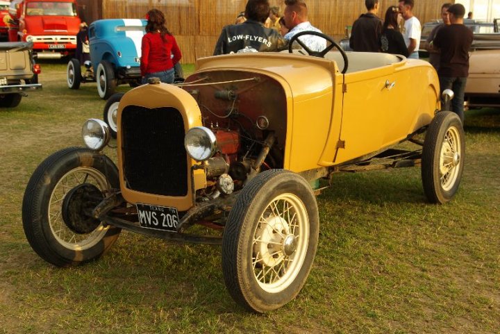 Pistonheads - The image captures a scene on a grassy field, dominated by a classic yellow truck with its hood propped open, revealing the engine. The truck, with its vintage design and sturdy appearance, is positioned in the center of the frame, drawing the viewer's attention. A group of people, dressed casually, are scattered around the area, mingling in a relaxed manner. In the background, there are additional vehicles, including a red truck and a blue car, hinting at a larger event or gathering. The atmosphere seems casual and leisurely, with the open field serving as a temporary parking lot for these vehicles.