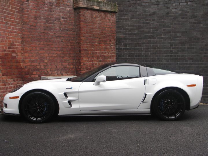 How about some ZR-1 photos to cheer you up? - Page 1 - Corvettes - PistonHeads - The image features a white sports car parked against a brick wall and a black wall. The car appears to be a convertible with a sleek design, with a black soft-top and black accents on the body. Its black rims and tires contribute to its sporty aesthetic. The vehicle is positioned facing forward with the top up, which suggests it might be ready for use or display. The car's position in front of the brick wall adds an urban and somewhat rugged feel to the image.