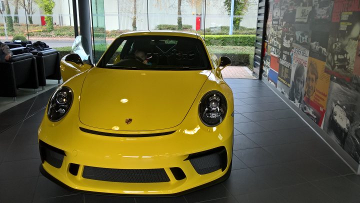 991.2 GT3 - Colours. Spec. Q+A. etc etc.. - Page 287 - 911/Carrera GT - PistonHeads - This is a photograph of a vibrant yellow Porsche sports car on display inside a building with a modern architectural design. The Porsche vehicle is positioned at a slight angle, showcasing its well-defined body panels, contours, and aerodynamic features. It is equipped with road tyres and features the brand's signature emblem on the nose. The background includes a variety of advertisements and images on the walls, suggesting an exhibition or showroom setting.