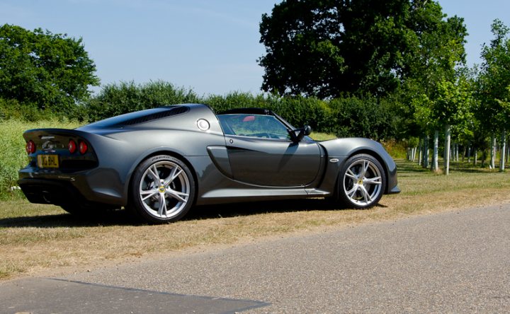 Exige Roadster  - Page 1 - Elise/Exige/Europa/340R - PistonHeads - The image presents a scene on a street lined with trees. A sports car is the primary focus, parked on the side of the road. The car has a sleek design with a black exterior and silver detailing, including a yellow stripe. It features big, shiny wheels that match the overall sporty aesthetic of the vehicle. The car is parked next to a strip of dirt on the grassy area, suggesting a park or a recreational area. The background of the scene is filled with lush green trees, providing a natural but serene backdrop to the image.