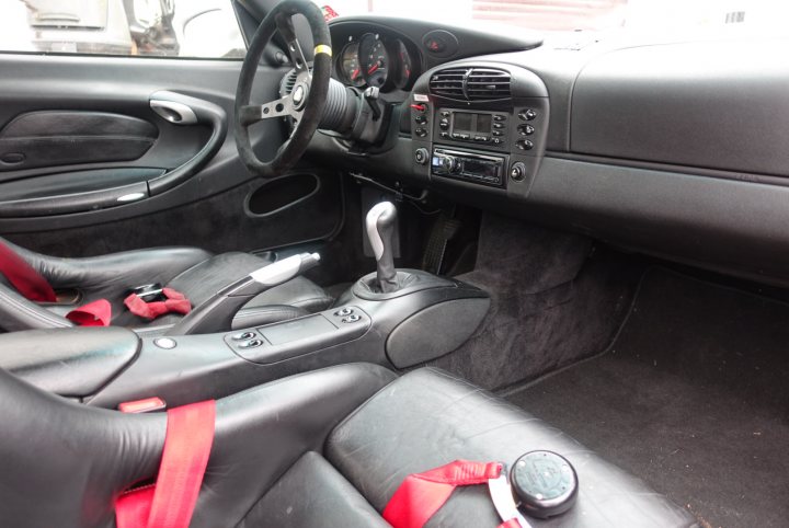 Porsche 996 interior design, time to see it in a new light? - Page 1 - 911/Carrera GT - PistonHeads - The image shows the interior of a car, specifically focusing on a pair of red seat belts extending from the car seat towards the gear shift. The black car seat is visible in the foreground, with the seat belt buckle already closed. The gear shift lever, located below the seat belt, appears to be in the middle of its range of motion. The car's dashboard is also visible in the background, featuring various control knobs and buttons. The lighting in the image is bright and even, indicating an open sunroof above.