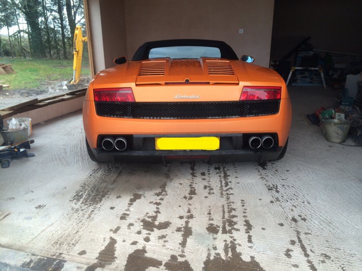 More pics of my LP560 - Page 1 - Gallardo/Huracan - PistonHeads - The image shows an orange sports car parked on a concrete driveway. The car has a sleek and aerodynamic design typical of sports cars, with a prominent rear wing and a deep scoop on the bonnet above the engine bay. The driveway appears to be part of a larger, open outdoor area with access to a garage or a workshop in the background. A small amount of oil or fluids can be seen on the ground next to the rear driver's side of the vehicle, suggesting some recent mechanical work or a routine check.