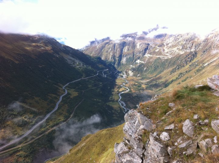 Alps Wheres Dam Pistonheads Hamster - This is a panoramic aerial view of a rocky mountainous landscape from a cliff. The terrain is rugged, with steep rocky faces and sinuous river valleys snaking between majestic peaks. The rocky surface provides a contrasting texture to the patchy green vegetation sporadically scattered across the valleys. A curving mountain road is visible, tracing a path along the side of the canyon. The bottom of the image offers a glimpse of a dense forest, adding another layer to the complex landscape. The sky above is clear, suggesting a bright day, and the overall scene conveys a sense of isolation and natural beauty.